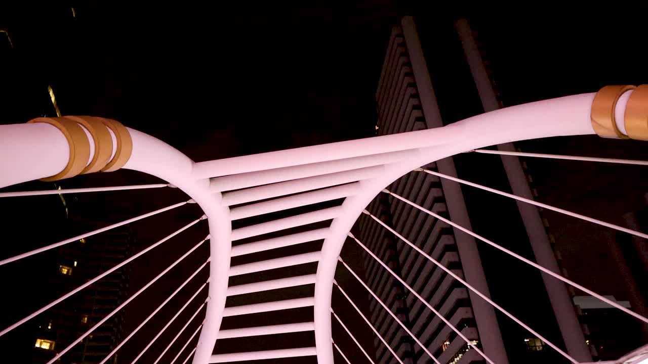 Man walks beneath brightly lit modern bridge at night, wide low-angle shot, urban environment