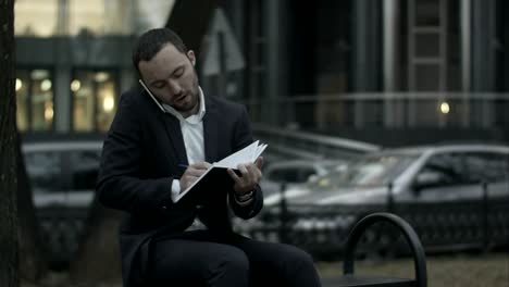 young bearded businessman talking by phone and writing in diary, working with notes at green park