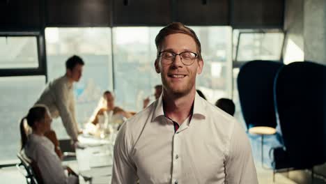 Ein-Blonder-Mann-Mit-Brille-Und-Weißem-Hemd-Posiert-Im-Büro-Vor-Dem-Hintergrund-Seiner-Golegs,-Die-An-Einem-Tisch-In-Einem-Büro-Mit-Panoramafenstern-Arbeiten