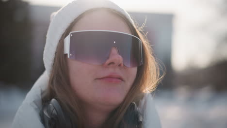 young woman adjusting tinted goggles over white beanie with blissful smile and wind and blown hair against snowy urban skyline under bright winter sunshine glinting off frosted streets