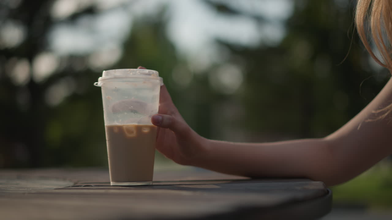 Donna prende un caffè freddo con ghiaccio nel patio, pausa di una pendolare in un bar sul ciglio della strada con luce solare laterale, gesto casuale del braccio, bicchiere di plastica da asporto con condensa, atmosfera estiva spontanea in città e ritmo rilassato