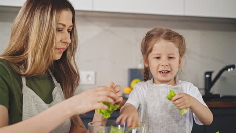 Mujer-Con-Hija-Divertida-Lágrima-Hojas-De-Lechuga-Cocinando-Ensalada