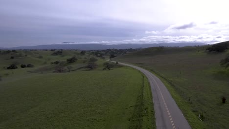 Aerial-shot-of-rural-road-with-no-cars-weaving-its-way-through-lush-green-hills