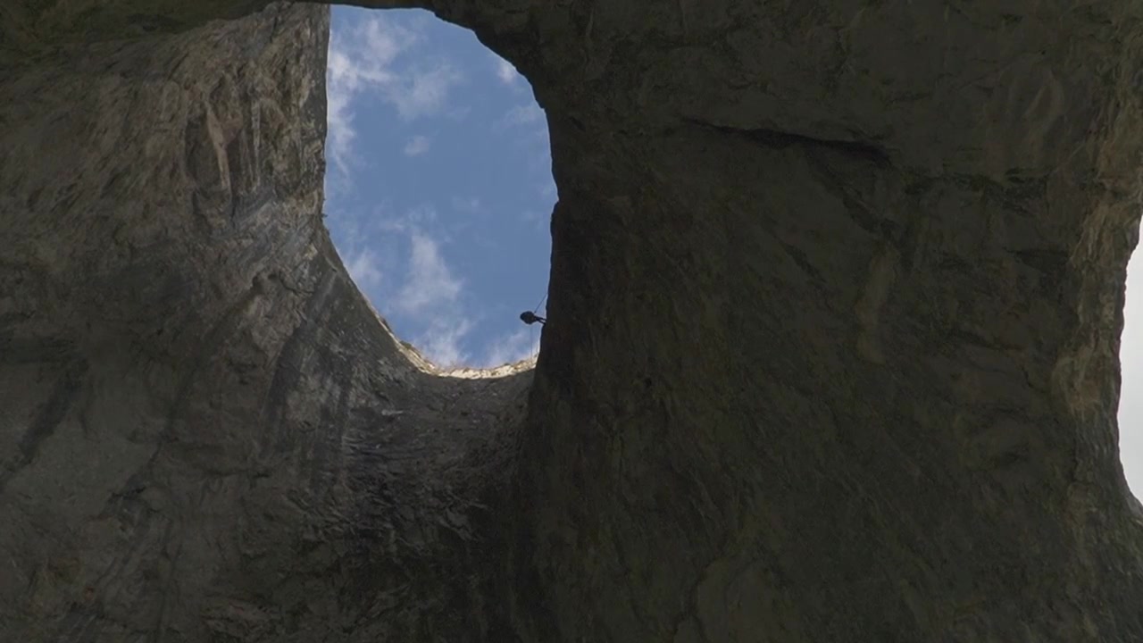 Steady low angle shot looking up at a small silhouette of a climber starting a rope descent from the edge of the massive Coșul Boului rock arch in Romania, against a cloudy sky.