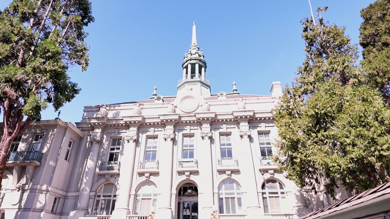 Vista da histórica Antiga Câmara Municipal em Berkeley, Califórnia, mostrando sua bela arquitetura Beaux-Arts com uma torre de relógio central e altas árvores verdes emoldurando o edifício