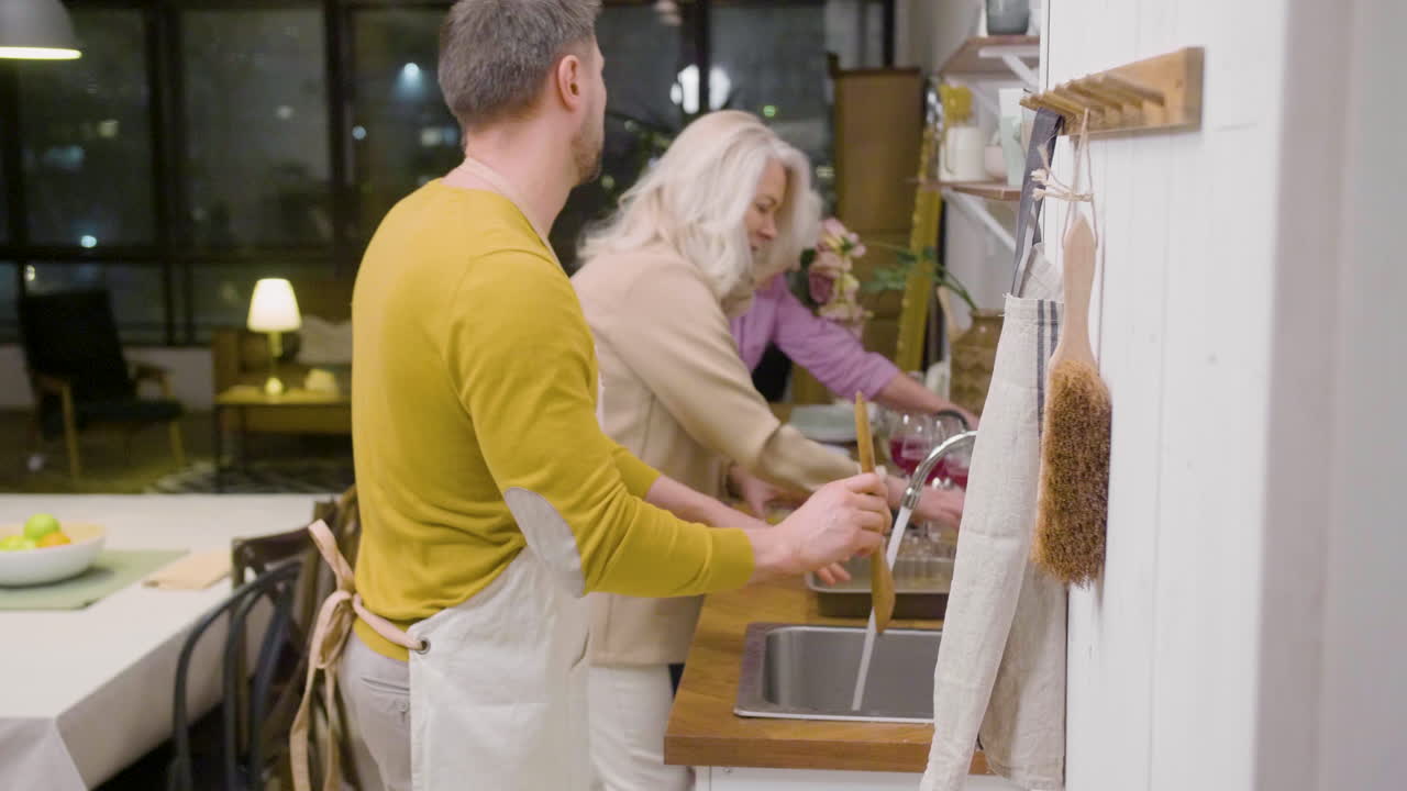 Side View Of A Man Washing The Family Dinner Dishes At The Sink In The Kitchen While A Mature Woman Helping Him