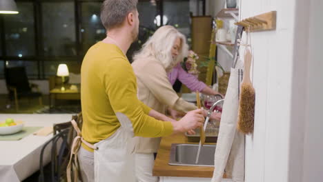 side view of a man washing the family dinner dishes at the sink in the kitchen while a mature woman helping him