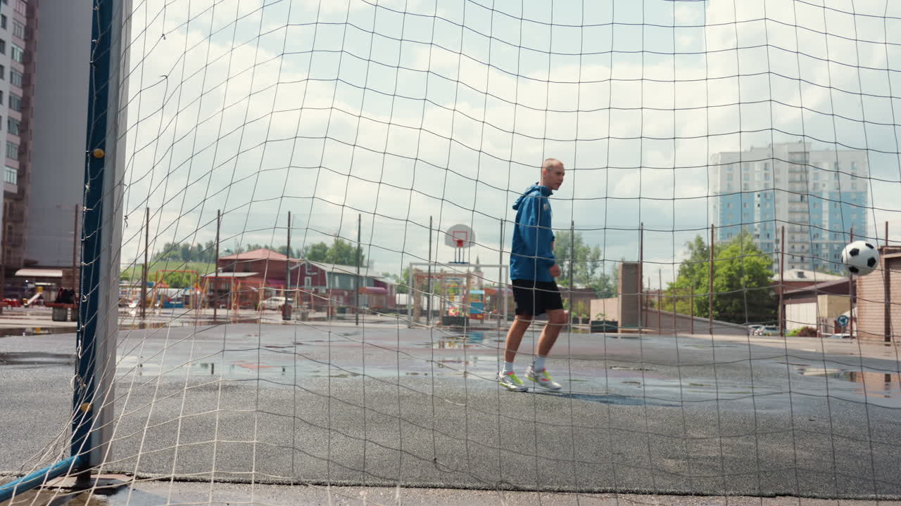 goalkeeper training on wet urban pitch, approaches net and prepares to kick, boots splash through puddles, apartment towers rise in background, overcast sky casts soft light, gritty cinematic mood
