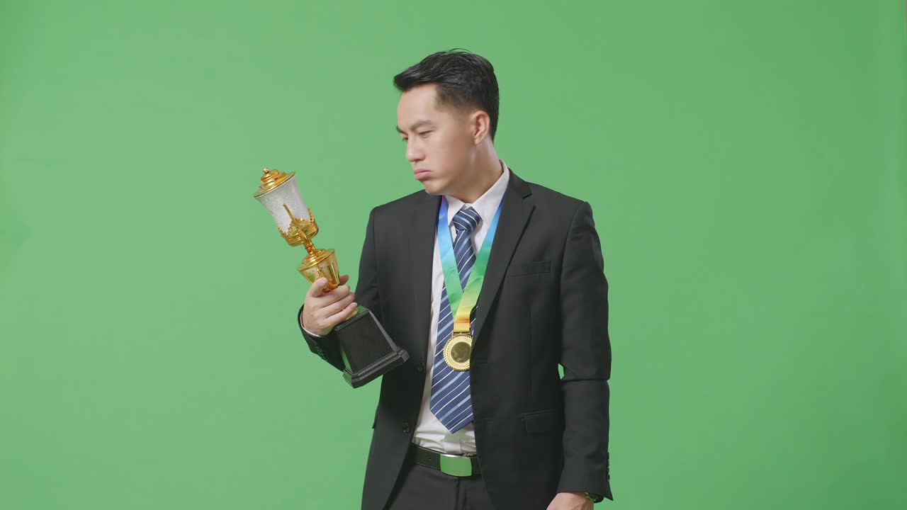 슈트와 타이를 입고 금메달을  아시아 사업가, 트로피를 쳐다보며 머리를 흔들며 불만을 품고, 스튜디오의 녹색 스크린 배경에서 첫 번째 우승자로 승리합니다.