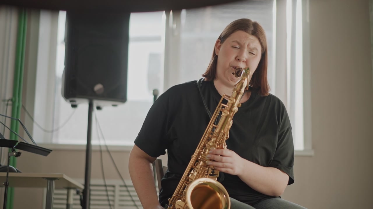 Sassofonista in un piccolo studio di pratica, l'insegnante di musica dimostra un tono caldo e il controllo del respiro durante una lezione privata, microfono e altoparlanti sullo sfondo, luce naturale soffusa dalla finestra, atmosfera rilassata.