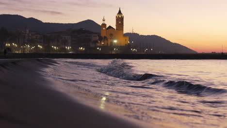 coastal sunset skyline is reflected in sea waves washing ashore, silhouette of church stands against backdrop of majestic mountain range