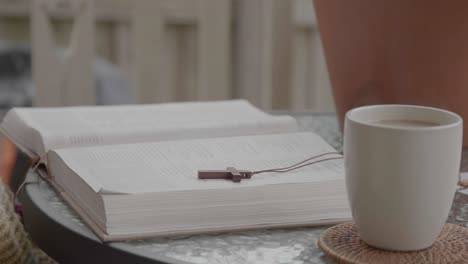 open bible with a wooden cross necklace laid on its pages, hot coffee mug on table