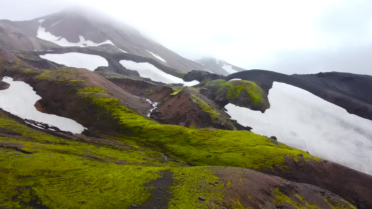 Aerial drone footage gliding and flying forward over green mossy and white snow civerd dark volcanic Iceland terrain that has a large mountain in fog in the background during cloudy day. Beautiful.