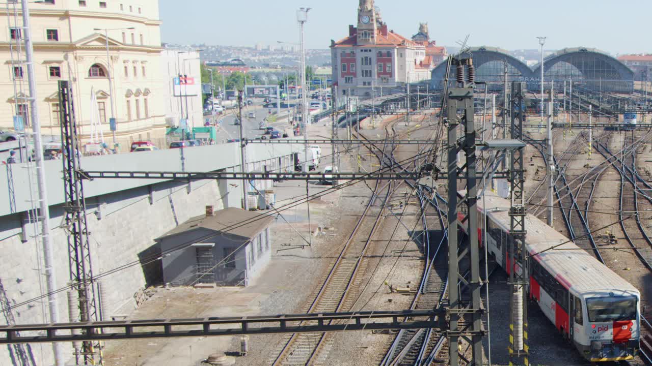 A red and white passenger train approaches the historic main railway station in Prague on a sunny day, captured from an elevated, wide-angle perspective