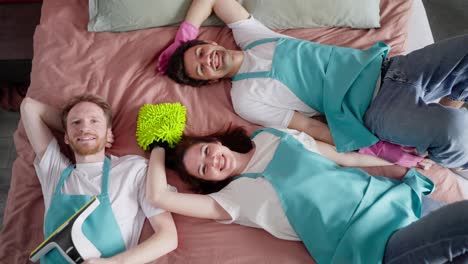 Portrait-of-a-trio-of-cleaners-in-white-t-shirts-and-blue-aprons-lying-on-a-bed-on-a-pink-blanket-after-heavy-cleaning-in-a-modern-apartment-and-bedroom