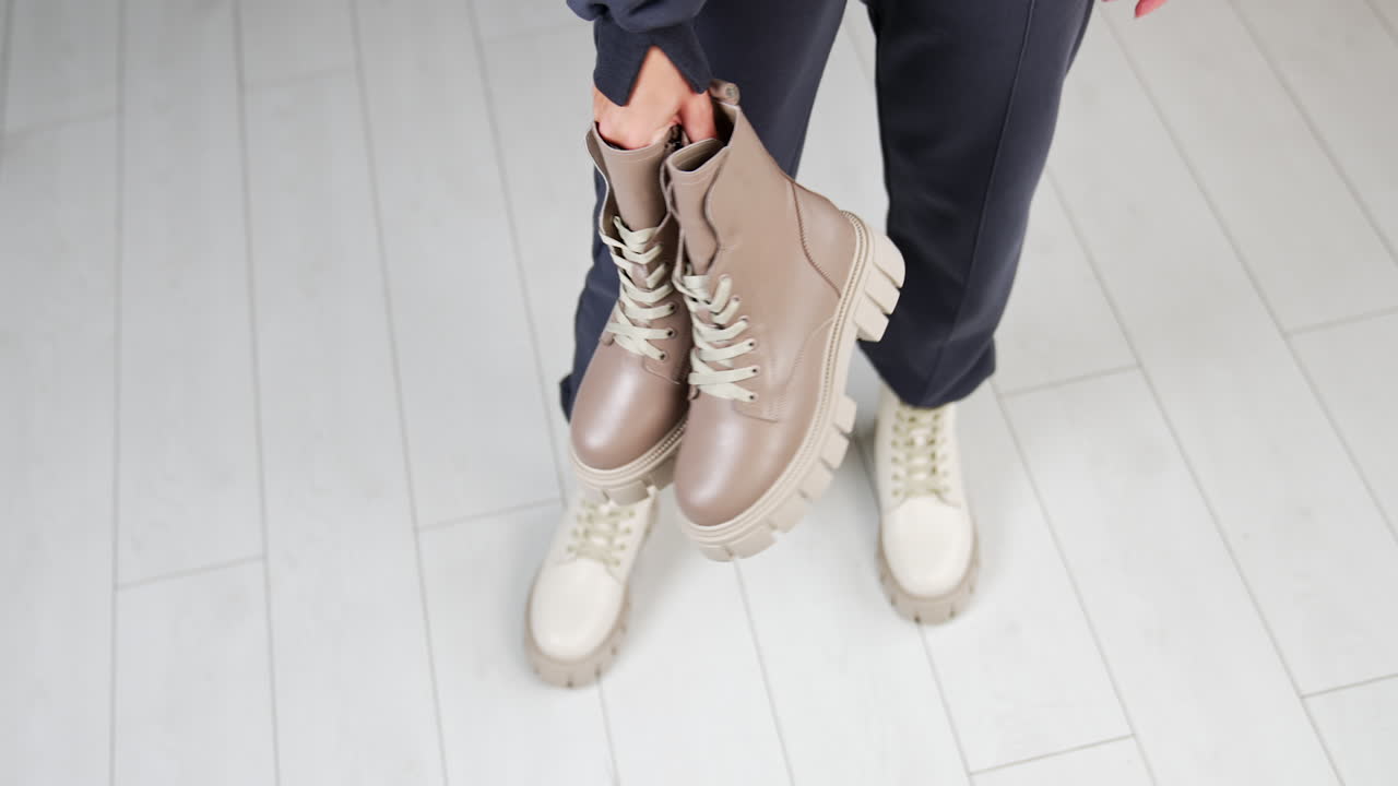 Female model wearing loose pants and white tractor sole boots holding another pair of beige boots in hands. View from above. White floor backdrop.