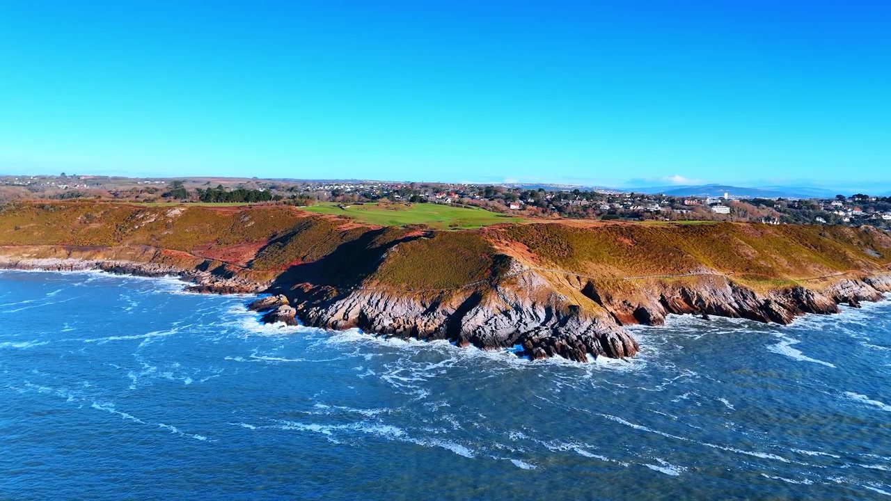 Vista Aérea de Ponta Costeira Rochosa com Ondas Quebrando e Aldeia. Ondas brancas quebram sobre uma ponta rochosa irregular enquanto uma aldeia na encosta e cabanas de praia margeiam a costa atrás dela