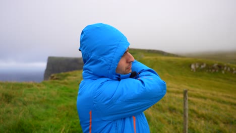 medium view of man covering head with blue jacket hood, walking along mykines coastline