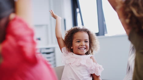 Infant-school-kids-with-hands-in-the-air-in-class,-learning-to-count-with-their-teacher,-close-up