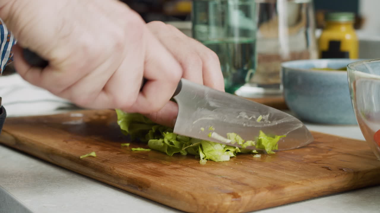primer plano de las manos del hombre cortando lechuga y agregándola en un tazón de vidrio para preparar una ensalada saludable