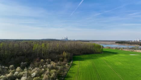 Kraftwerksstation-Mit-Rauchigem-Schornstein-In-Konin,-Polen-Drohnenflug-über-Klarem-Seewasser-Und-Grünem-Feld