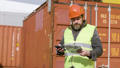 trabajador con chaleco y casco de seguridad leyendo documentos y usando un smartphone en un parque logístico