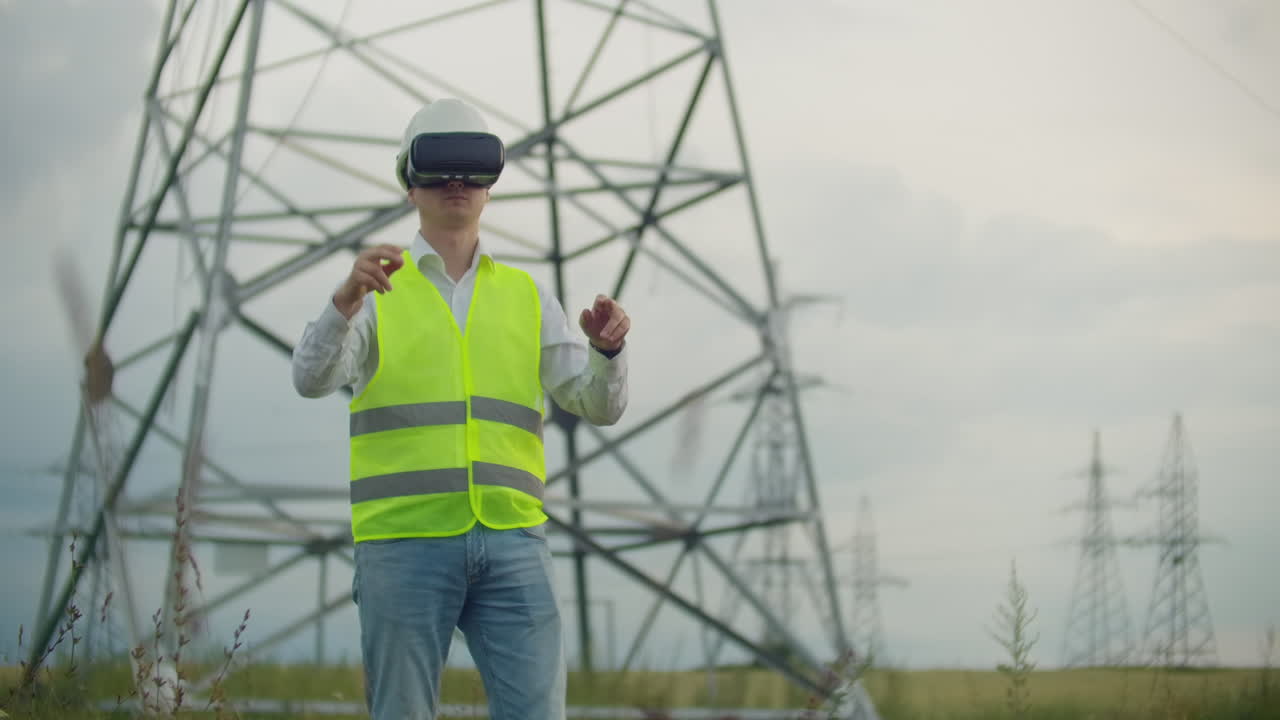 электрик-мужчина в очках виртуальной реальности движет рукой, имитируя работу с графическим интерфейсом электростанции на фоне высоковольтных электрических линий.