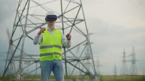 A-male-electrician-in-virtual-reality-glasses-moves-his-hand-simulating-the-work-with-the-graphical-interface-of-the-power-plant-against-the-background-of-high-voltage-electric-transmission-lines.