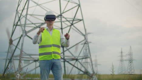 un electricista masculino con gafas de realidad virtual mueve su mano simulando el trabajo con la interfaz gráfica de la central eléctrica contra el fondo de líneas de transmisión eléctrica de alto voltaje.