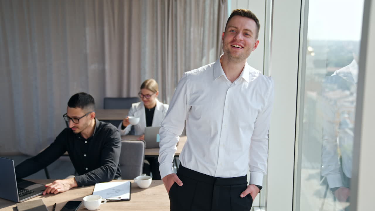 Happy confident handsome businessman standing near the panoramic window. People work at laptops at backdrop.