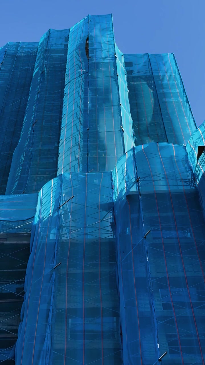 Malha azul envolve edifício alto em construção contra céu claro, vista ao nível da rua.