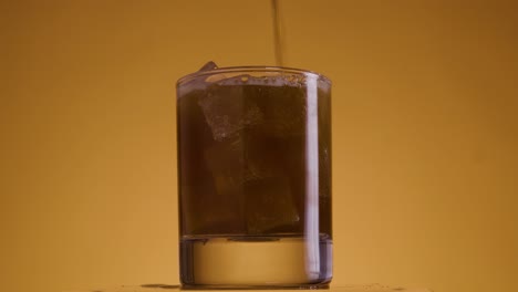 toma estática de vidrio lleno de cubitos de hielo contra un fondo amarillo dorado con un ajuste de luz naranja en el que se llena la cola