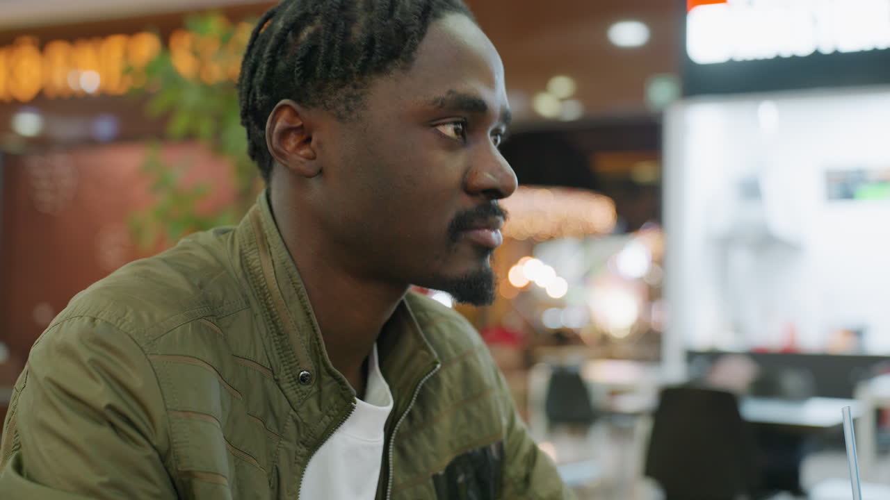 Close up of young man posing with warm smile resting hand under chin, wearing green jacket in indoor setting with blurred background lights, relaxed thoughtful mood