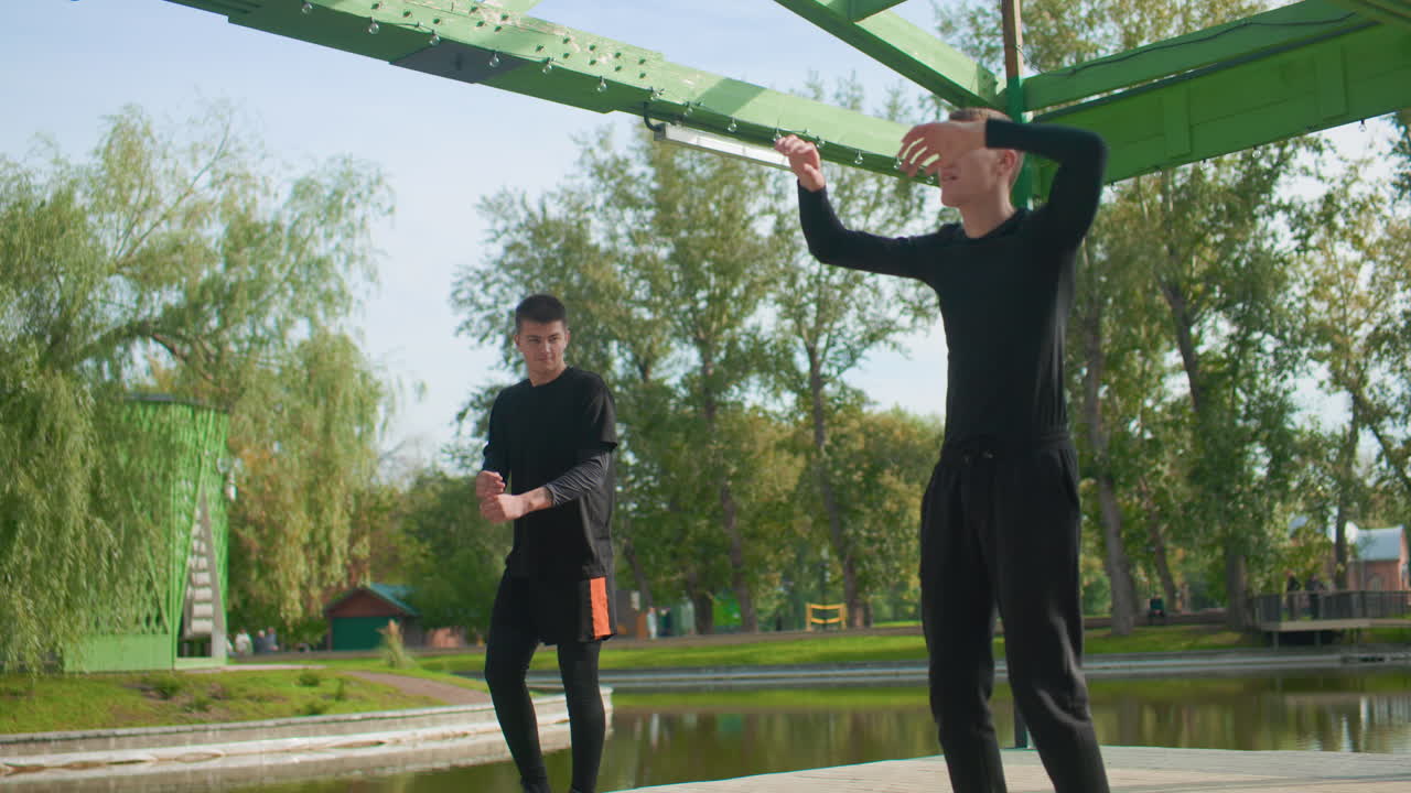 To hvide mænd øver udtryksfuld koreografi under en metalpavillon. De er i sort tøj, med synkroniserede bevægelser og skarp timing. Partnerøvelser og improvisation nær en dam, plettet sollys gennem træerne