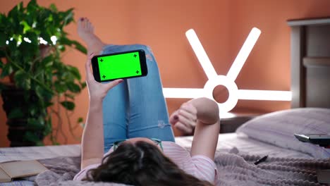 girl relaxing at home, lying on bed, holding modern smartphone with green screen. female swiping right and clicking waiting for result online. browsing internet concept
