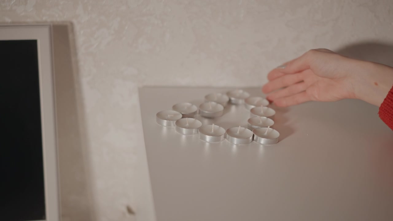 Close-up of hand with red knitted sleeve picking up and arranging cluster of round pills on white tabletop next to shelf; fingers grasping tablets indicate sorting or dosing action, soft indoor