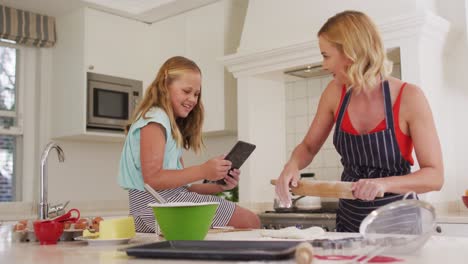 caucasian mother and daughter using digital tablet and baking together in the kitchen at home