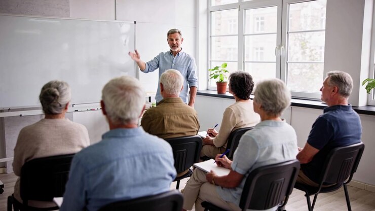 Extending right arm, instructor in blue shirt introducing lesson at classroom whiteboard, notebooks