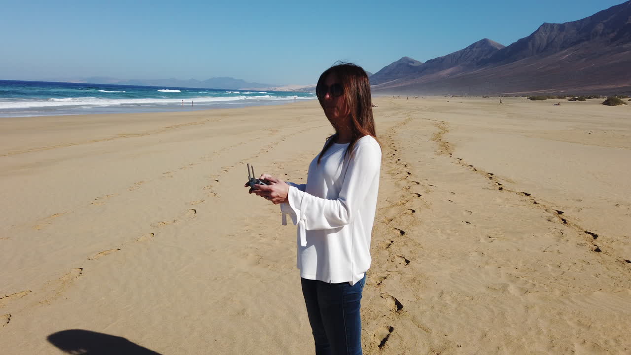 Woman operating a drone with a controller, standing on a sunny beach with the ocean behind