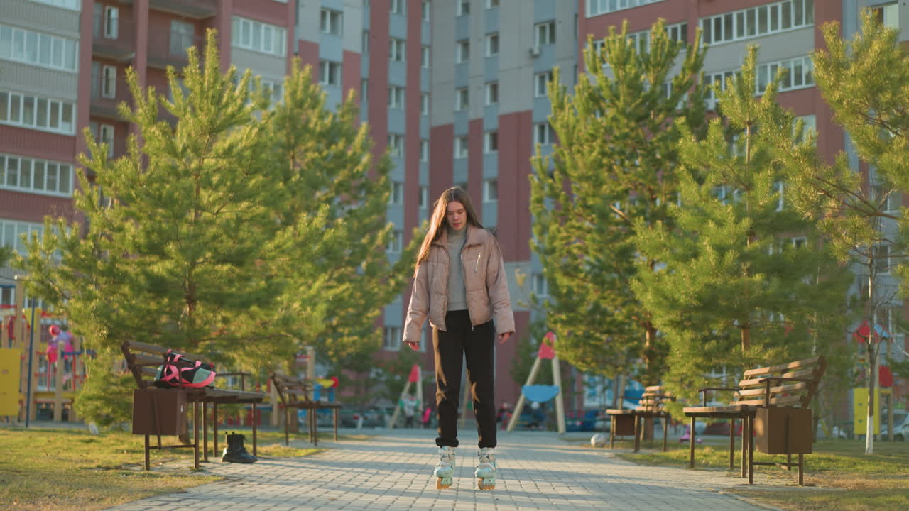 A young woman wearing a peach jacket and black trousers is rollerblading through a park lined with trees and benches. The urban setting features tall residential buildings in the background