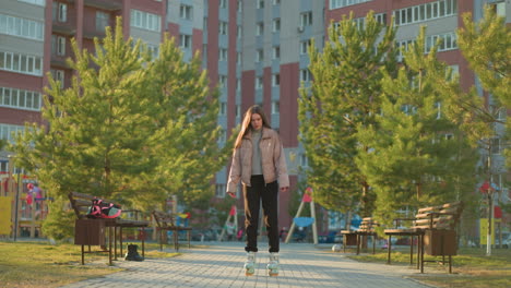 a young woman wearing a peach jacket and black trousers is rollerblading through a park lined with trees and benches. the urban setting features tall residential buildings in the background