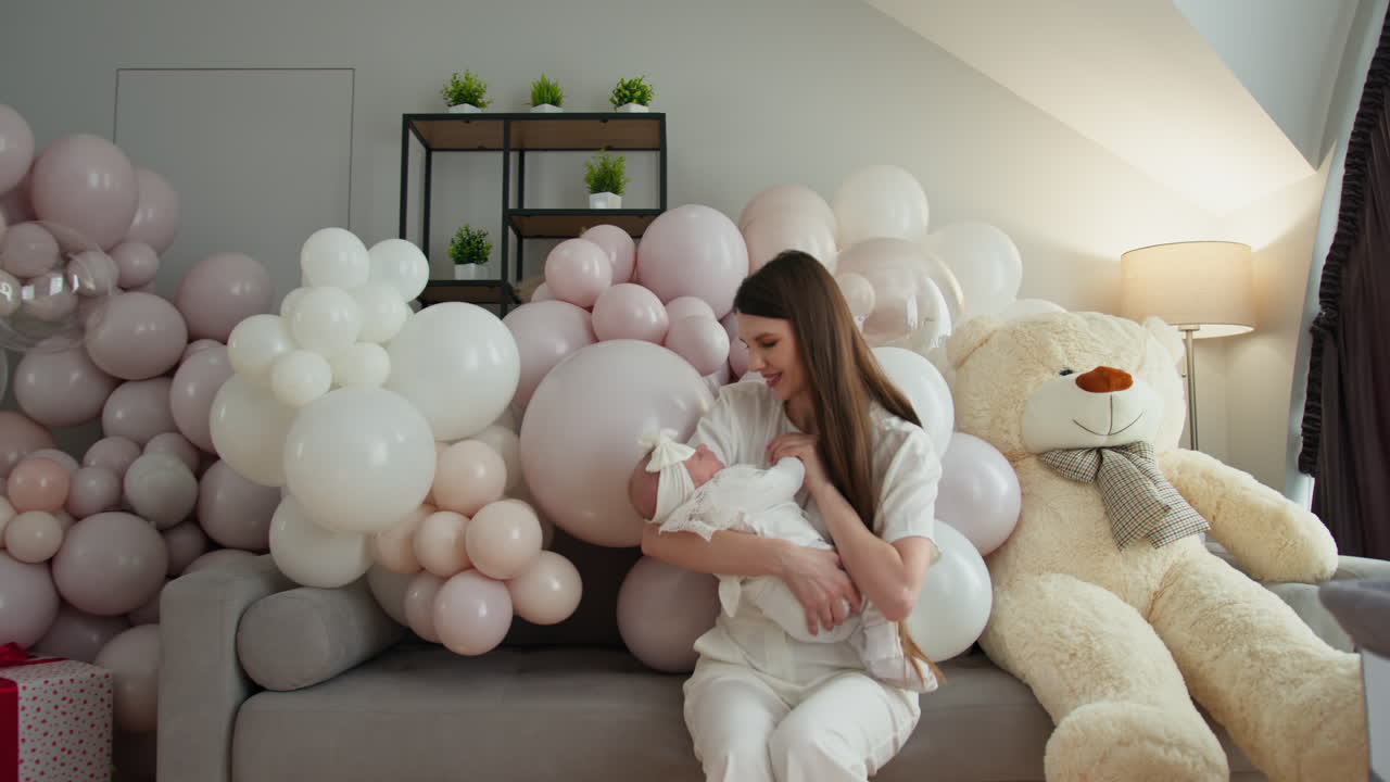 Madre sosteniendo a un bebé recién nacido rodeada de globos pastel y un osito de peluche celebrando la nueva vida en un ambiente acogedor.