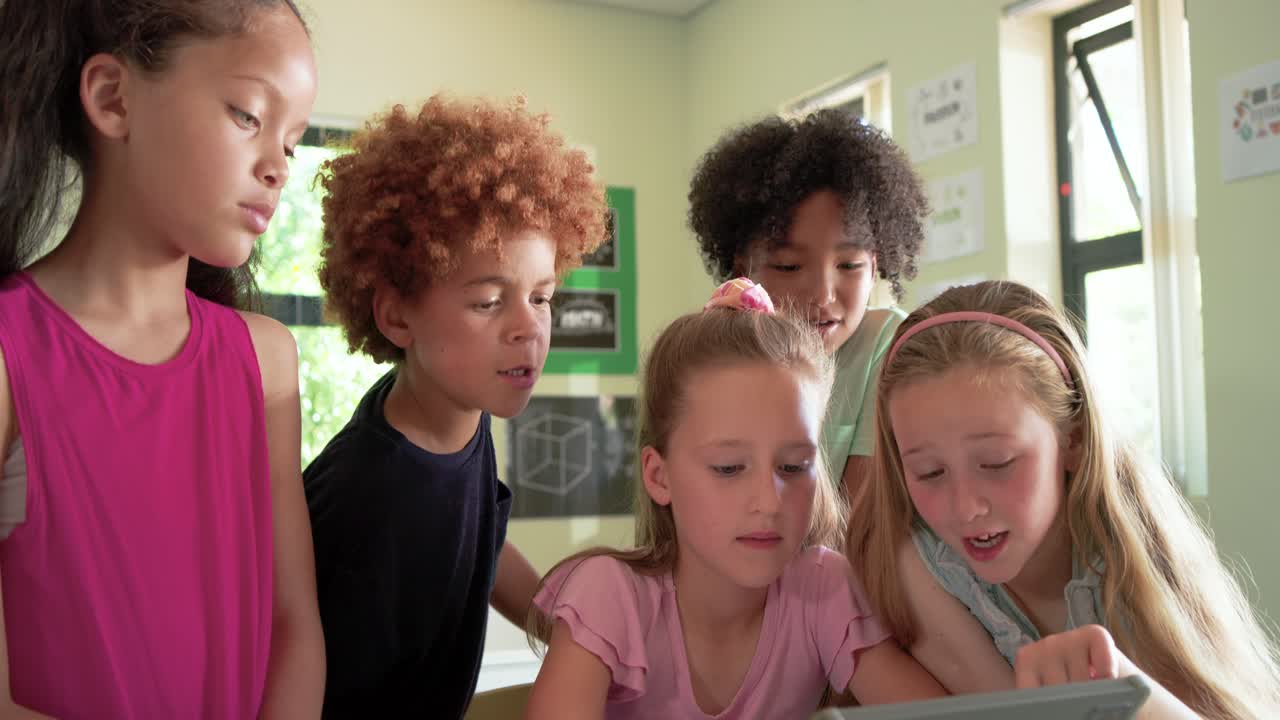 Schoolchildren leaning around tablet at classroom table, central girl touching screen to start game
