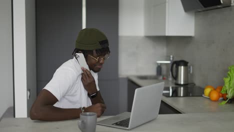Hombre-Negro-Trabajando-En-Casa-En-La-Cocina-Hablando-Por-Teléfono-Inteligente-Y-Usando-Una-Computadora-Portátil
