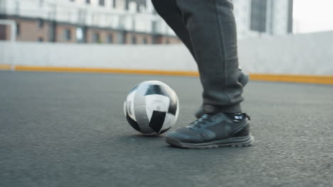 primer plano de un hombre controlando hábilmente una pelota de fútbol con los pies en la arena deportiva durante el entrenamiento, fondo borroso con edificios urbanos y goles