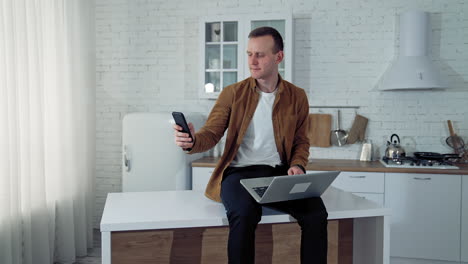 remote work from home. young man sits on the table with a laptop and using smartphone during the freelance job on the kitchen background.