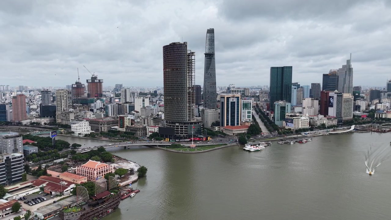 Yoğun bulutlu bir havada, Saygon Nehri boyunca 1. Bölge silüetine doğru ilerleyen bir drone çekimi; aşağıda Bitexco Finans Kulesi ve demirli nehir tekneleri, Ho Chi Minh Şehri, Vietnam.