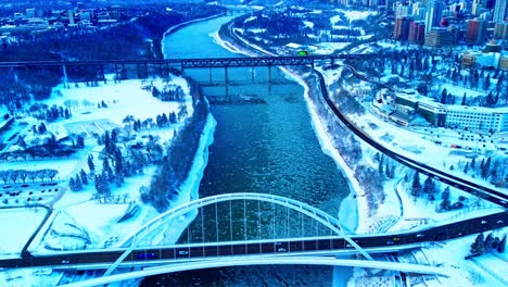 Bucle-Aéreo-De-Invierno-De-4k-Con-Vista-De-Pájaros-Sobre-El-Puente-Walter-Dale-Mientras-Los-Pedazos-De-Hielo-Fluyen-Por-El-Río-Saskatchewan-Norte,-Con-Cruces-De-Tráfico-Ligero-Hacia-El-Norte-Hasta-El-Centro-De-La-Ciudad-En-Medio-De-Los-Principales-Parques3-3