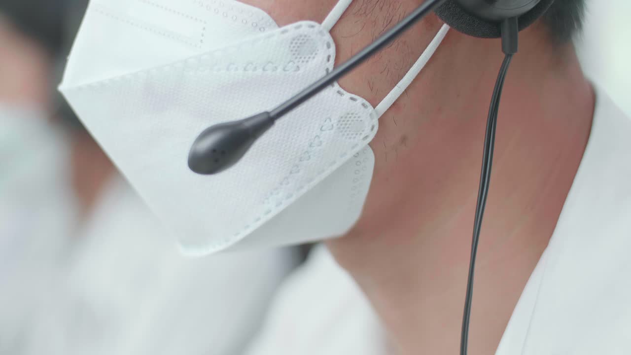 close up de un hombre asiático agente de centro de llamadas con auriculares y máscara hablando con el cliente en la llamada durante el trabajo con sus colegas en la oficina