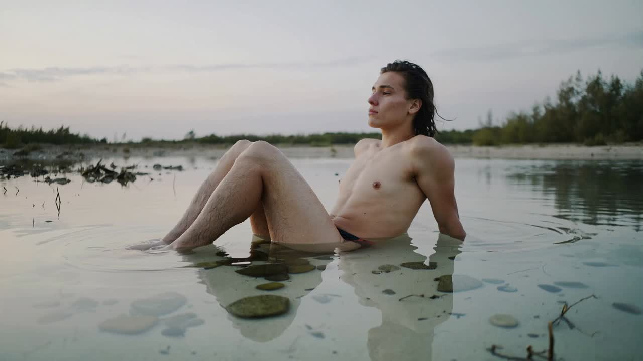 Sonnenuntergang beobachtend, zurückgelehnter Mann bewegt die Hand und hebt den Oberkörper im klaren Wasser, in dunkler Badehose.